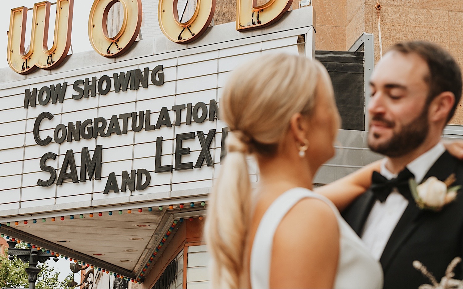 Close up photo of a bride and groom looking at one another, a movie theater is behind them with a sign saying "Now Showing Congratulations Sam and Lexi"
