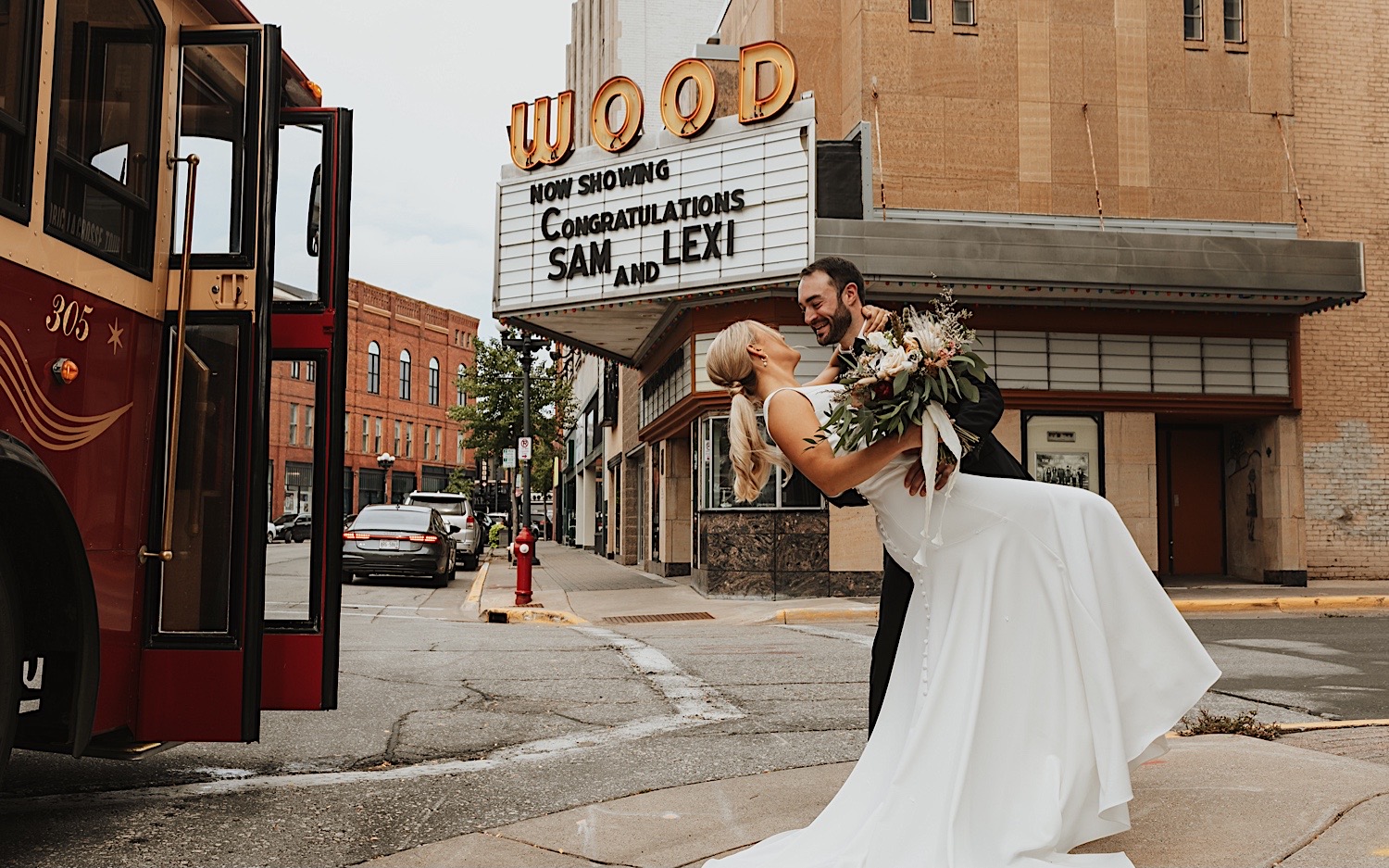 A bride and groom in downtown La Crosse smile as the groom dips the bride, a movie theater is behind them with a sign saying "Now Showing Congratulations Sam and Lexi"