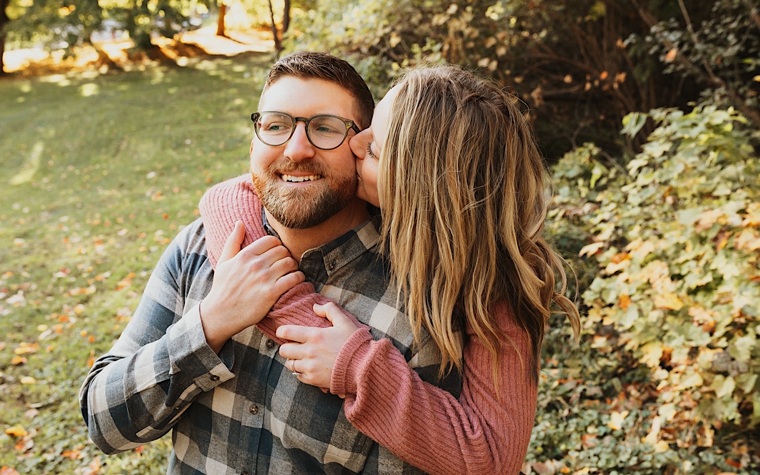 A man smiles as a woman hugs him from behind and kisses his cheek while the two are in a park with leaves on the ground during their fall engagement session in Winona