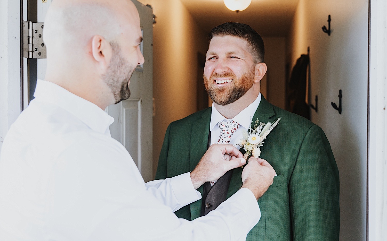 A groom smiles while standing in a doorway as a groomsman laughs while pinning on the groom's boutonniere 