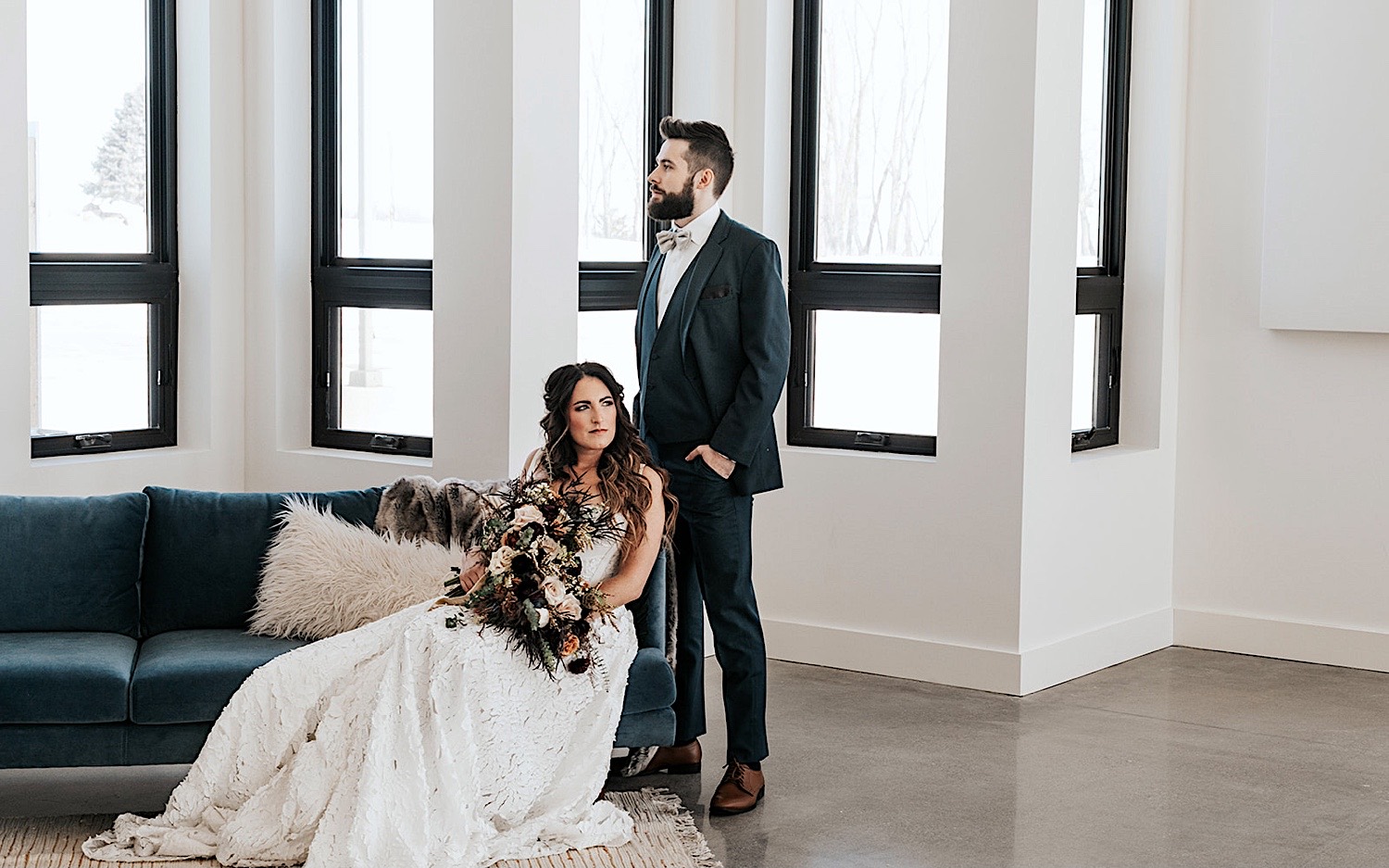 A bride sits on a couch while the groom stands next to her while inside of the naturally lit wedding venue, Woodhaven Weddings + Events