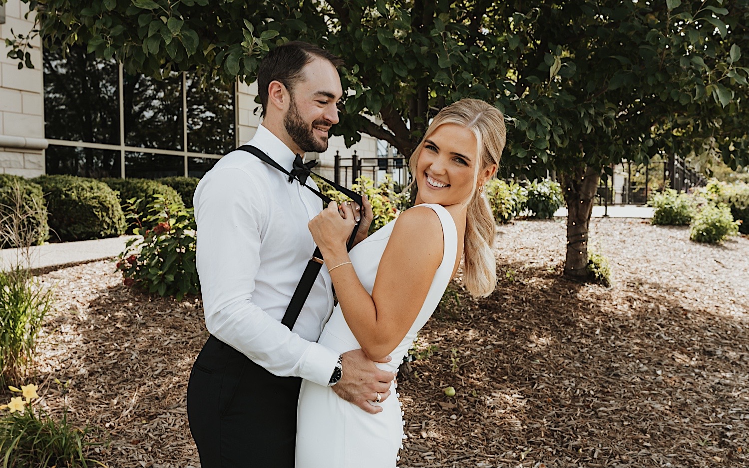 Portrait of a bride and groom, the bride is smiling at the camera and pulling on the groom's suspenders while he smiles at her