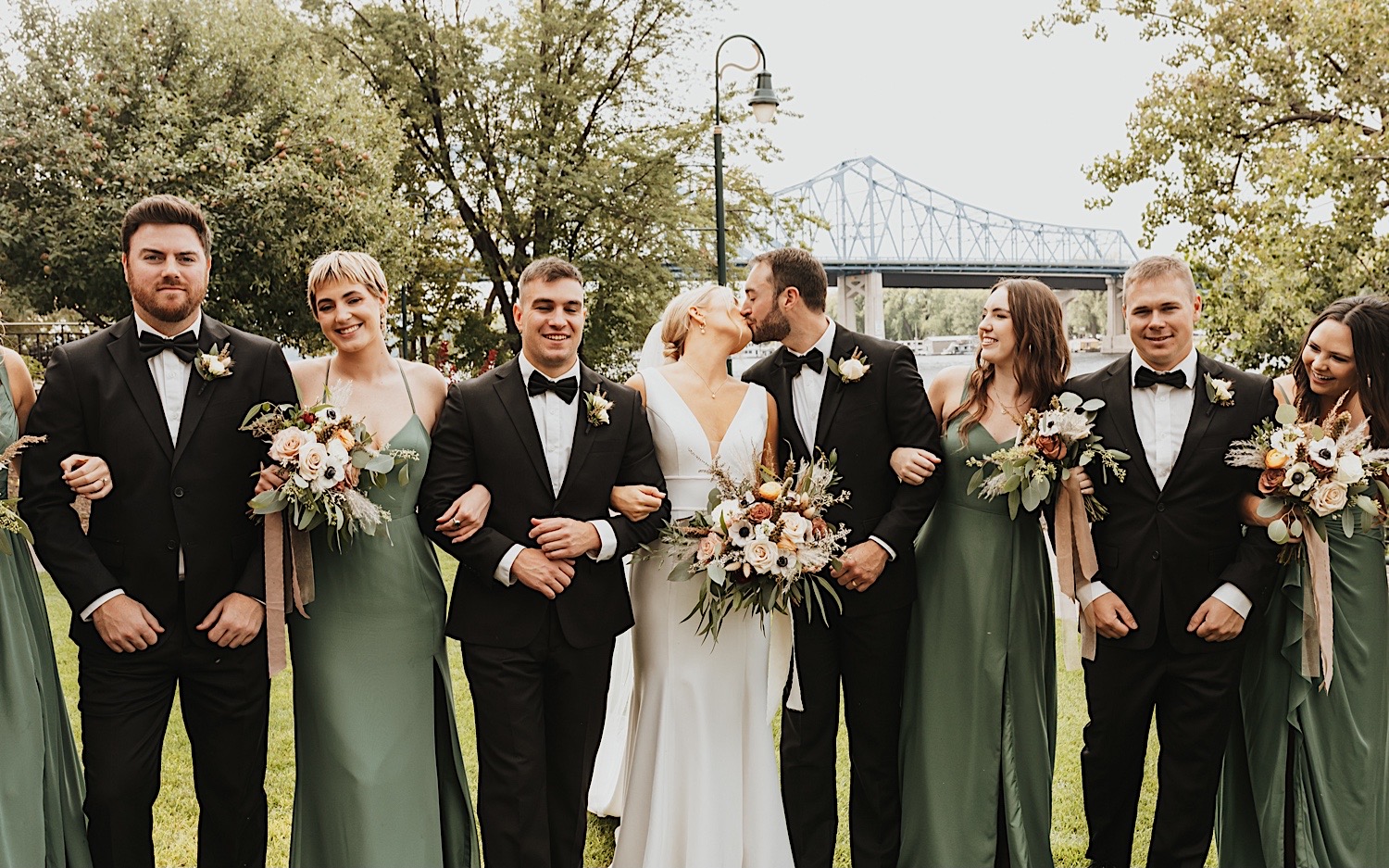 A bride and groom kiss while their arms are locked with their alternating wedding parties on either side of them