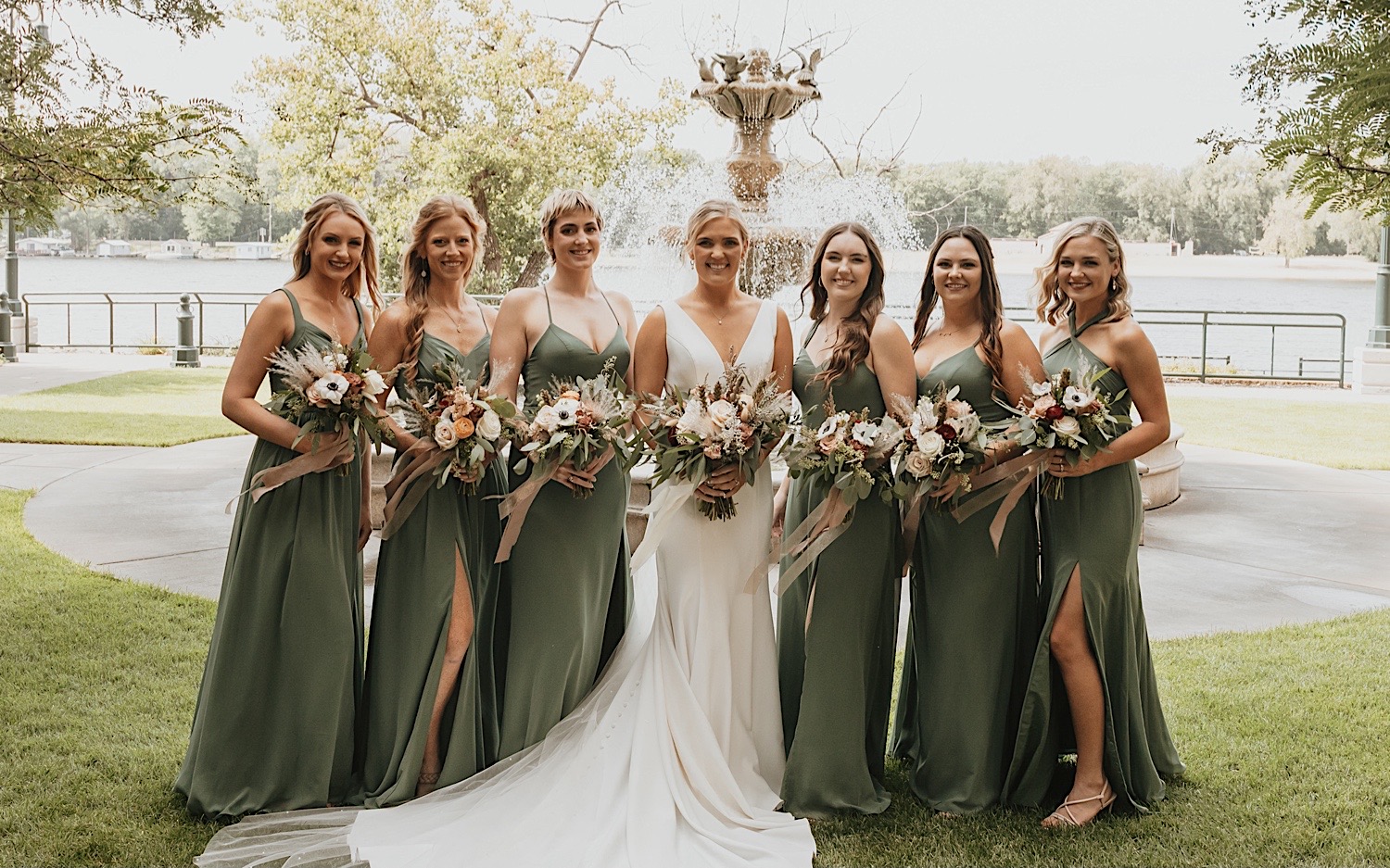 A bride and her bridesmaids smile at the camera while standing in front of a fountain
