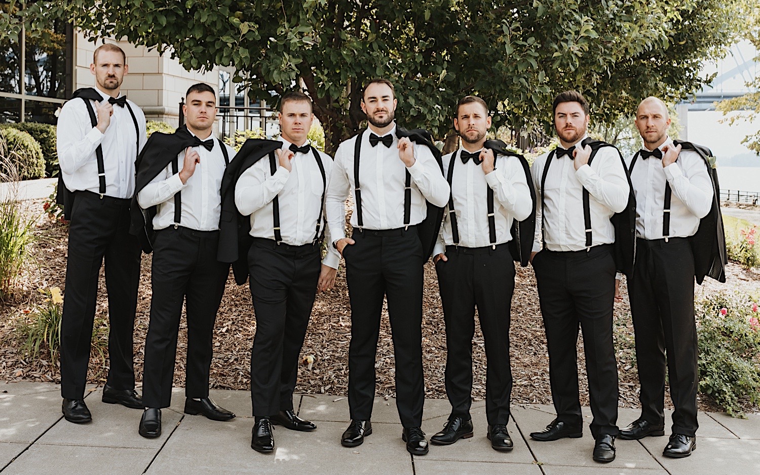 A groom and his groomsmen stand and look at the camera, each of them is holding their suit coat over one of their shoulders