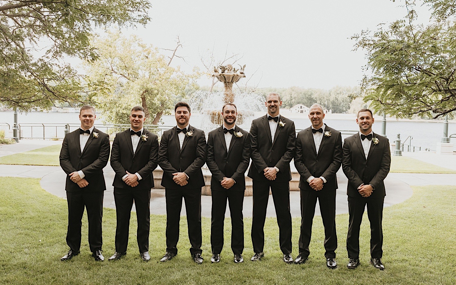 A groom and his groomsmen all smile and look at the camera while standing in front of a fountain