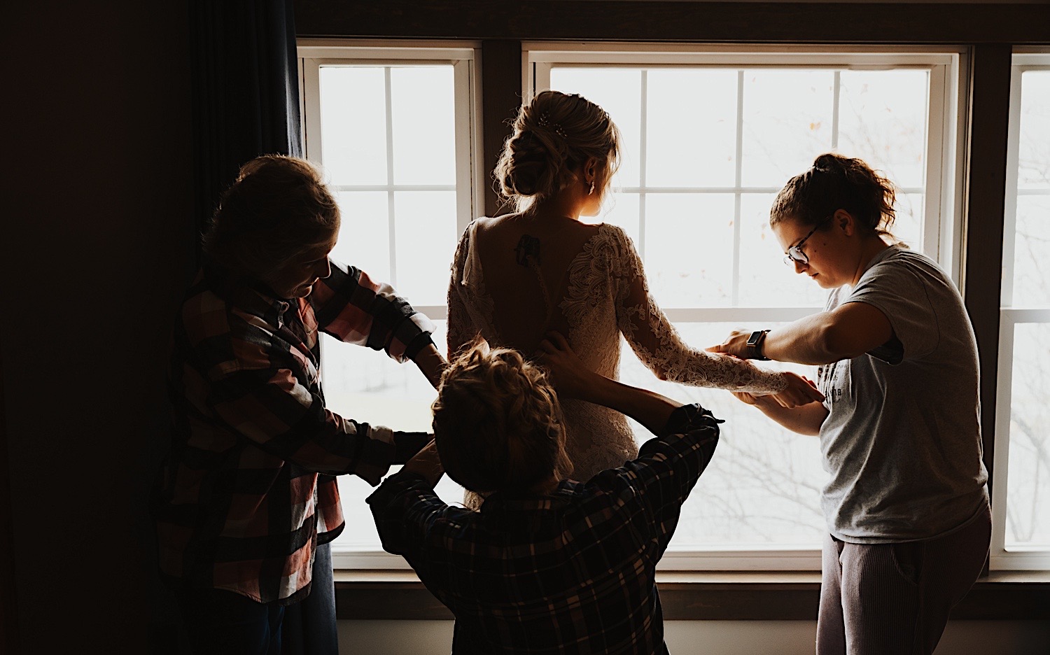 A bride looks out of a window while three women help to put on her wedding dress