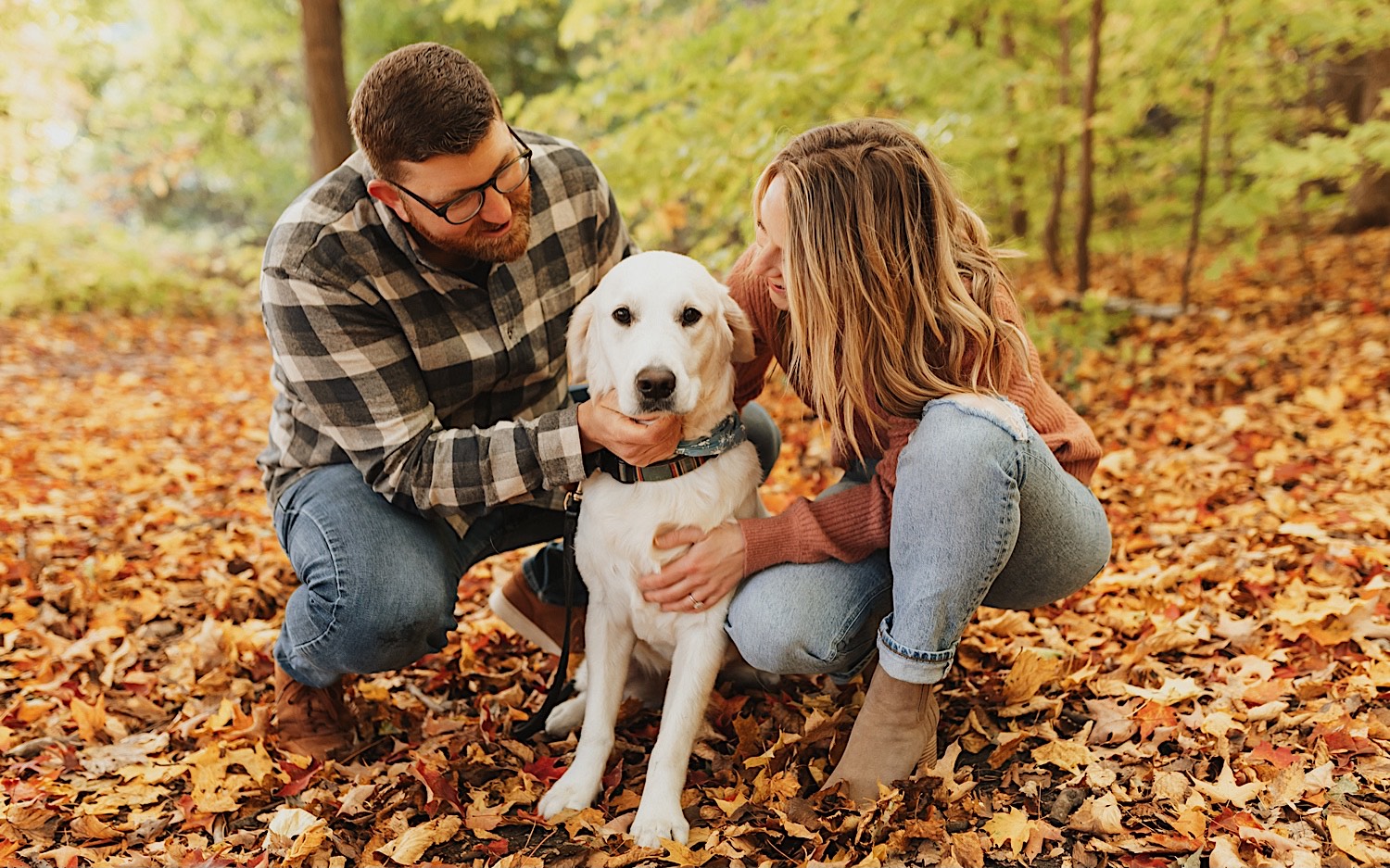 During their fall engagement session in Winona a couple squat down next to their dog who's sitting in between them and look at him while he looks at the camera, the ground is covered in leaves and trees are behind them 