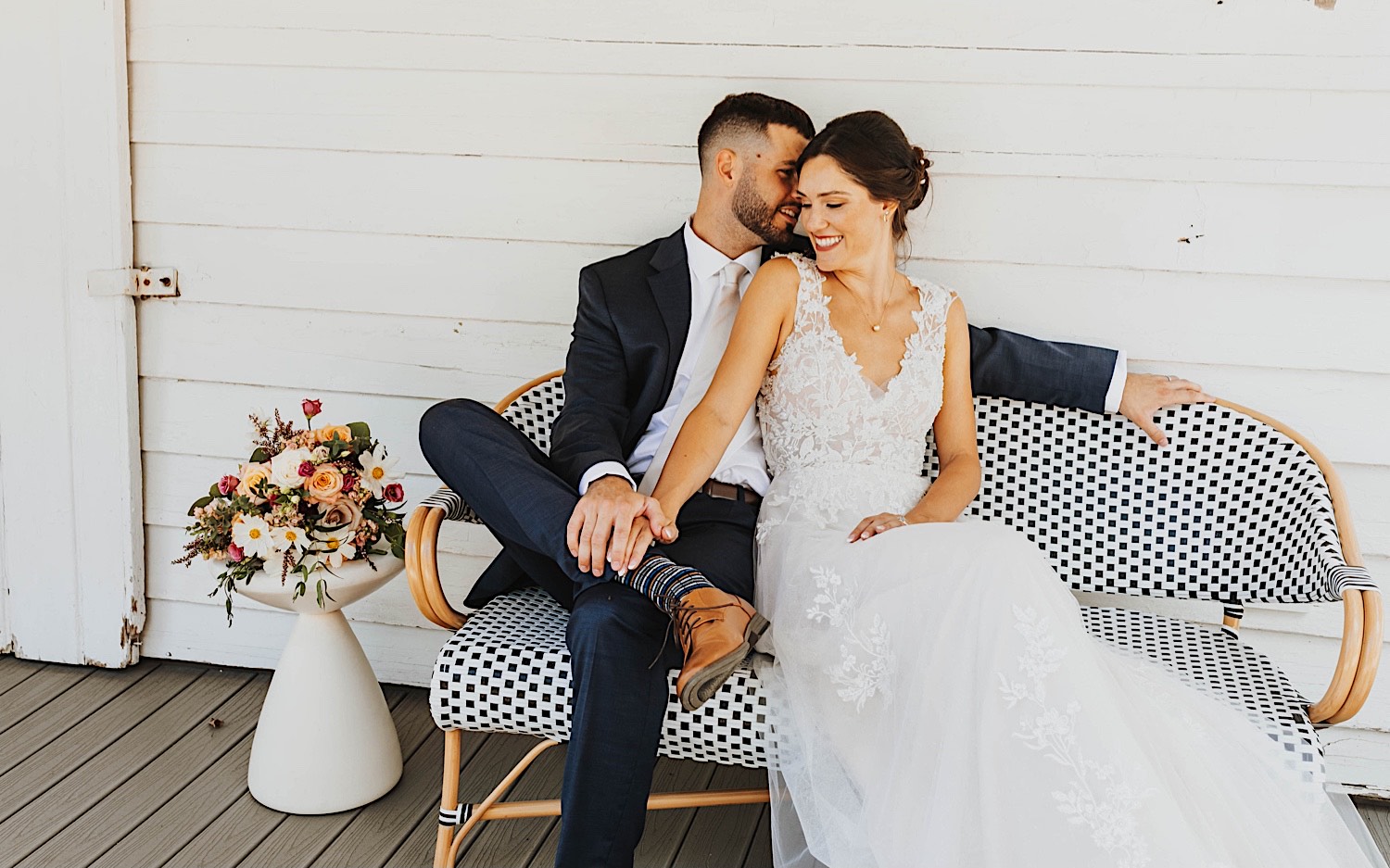 A bride and groom sit next to one another and smile on a bench outside of a house A bride and groom sit next to one another and smile on a bench outside of a house