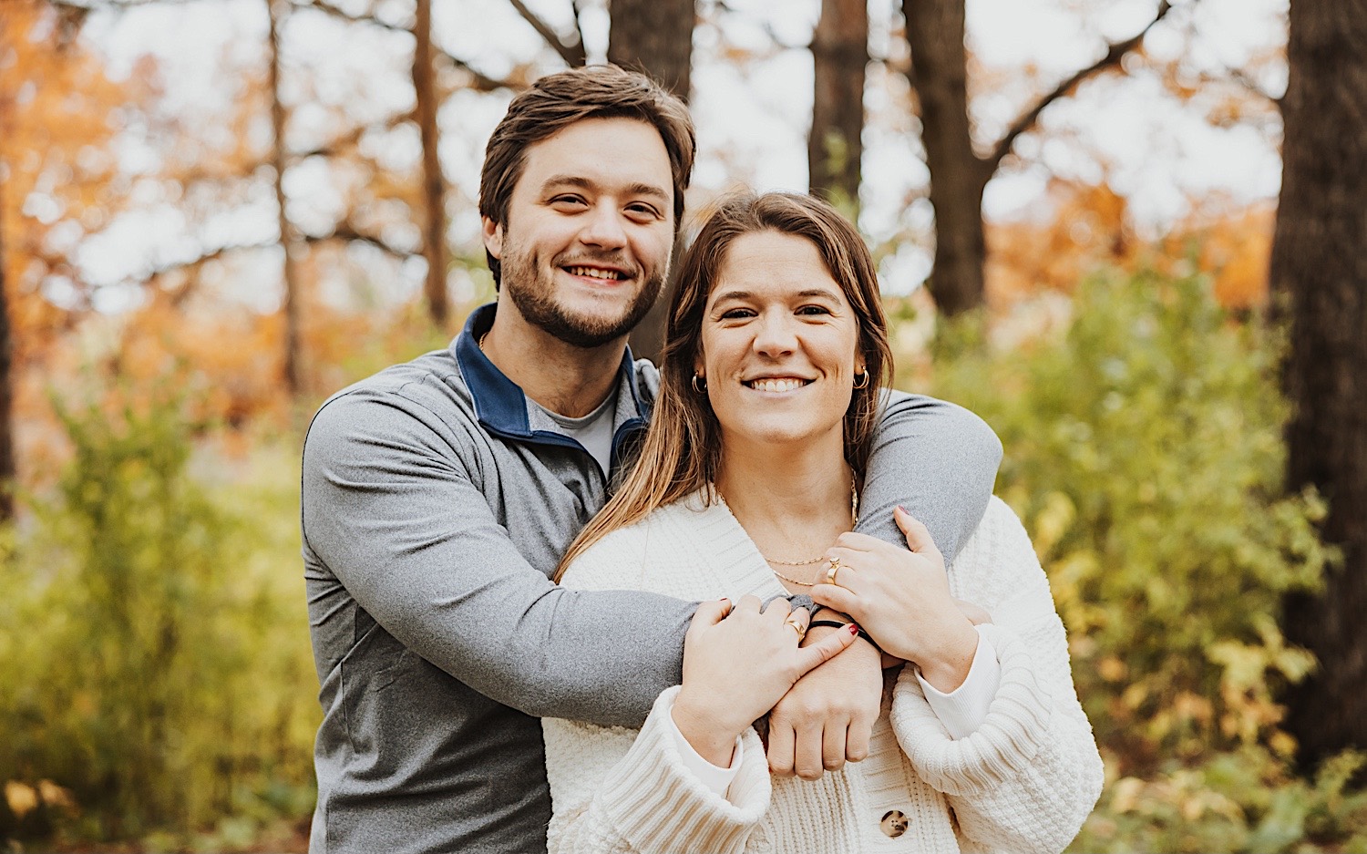 A man hugs a woman from behind as they both smile at the camera during their engagement session in a forest of Theodore Wirth Park in Minneapolis