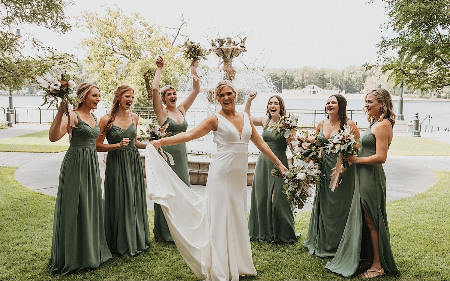 A bride smiles and plays with her dress as her bridesmaids cheer for her in front of a fountain
