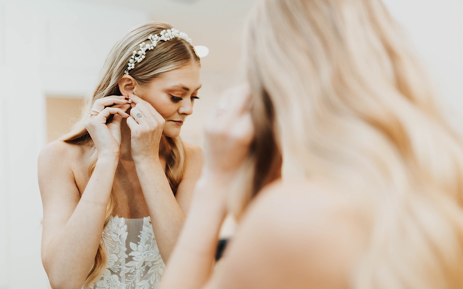 A bride in a mirror looks down as she puts on an earring as she gets ready for her wedding day at The Aisling