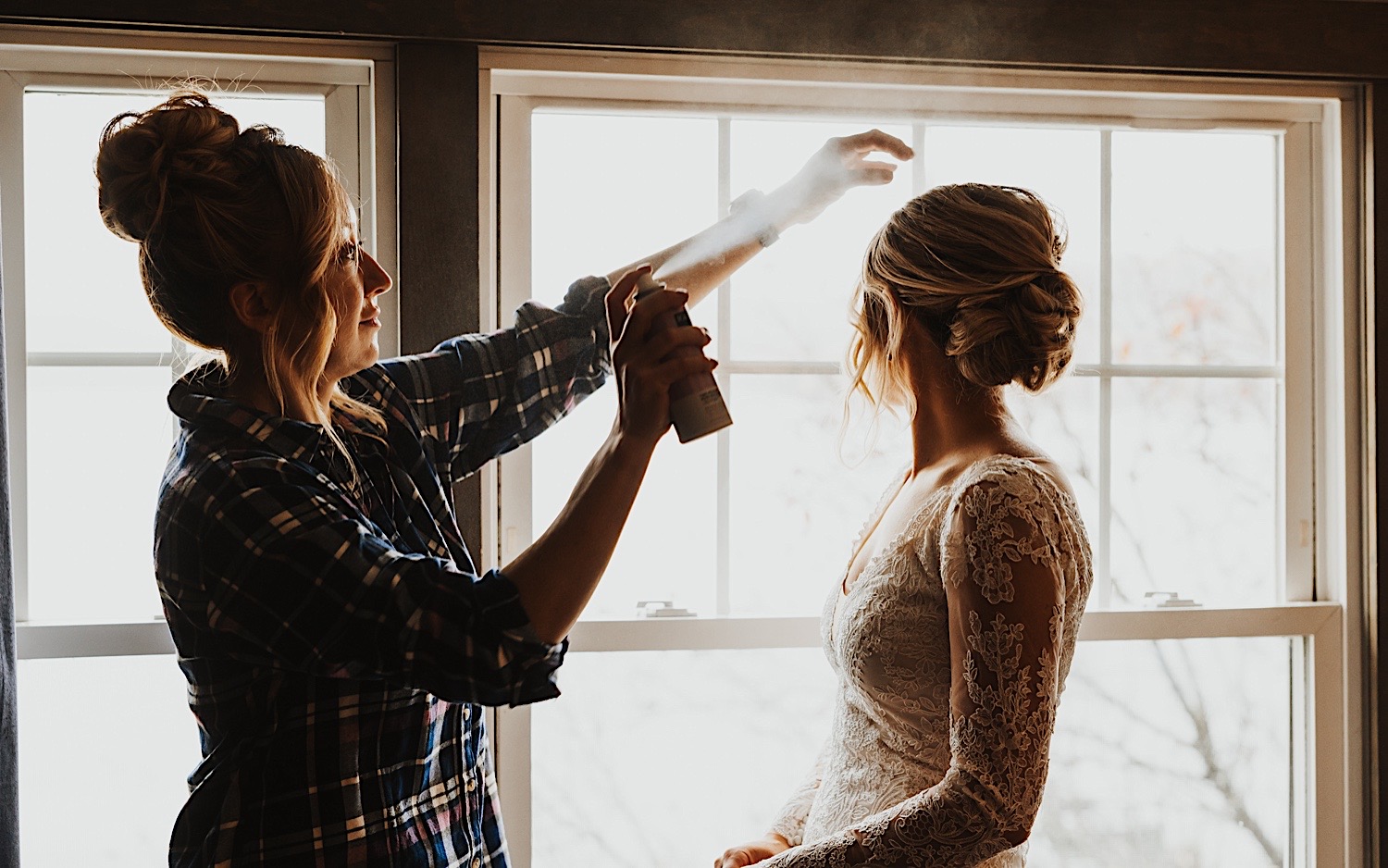 A bride looks to her right out of a window while a woman sprays hair spray in the air in front of the bride