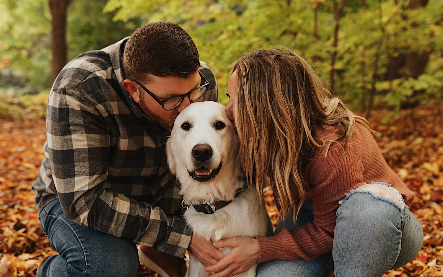 During their fall engagement session in Winona a couple squat down and kiss their dog who is sitting between them and is looking at the camera, the ground around them is covered in leaves