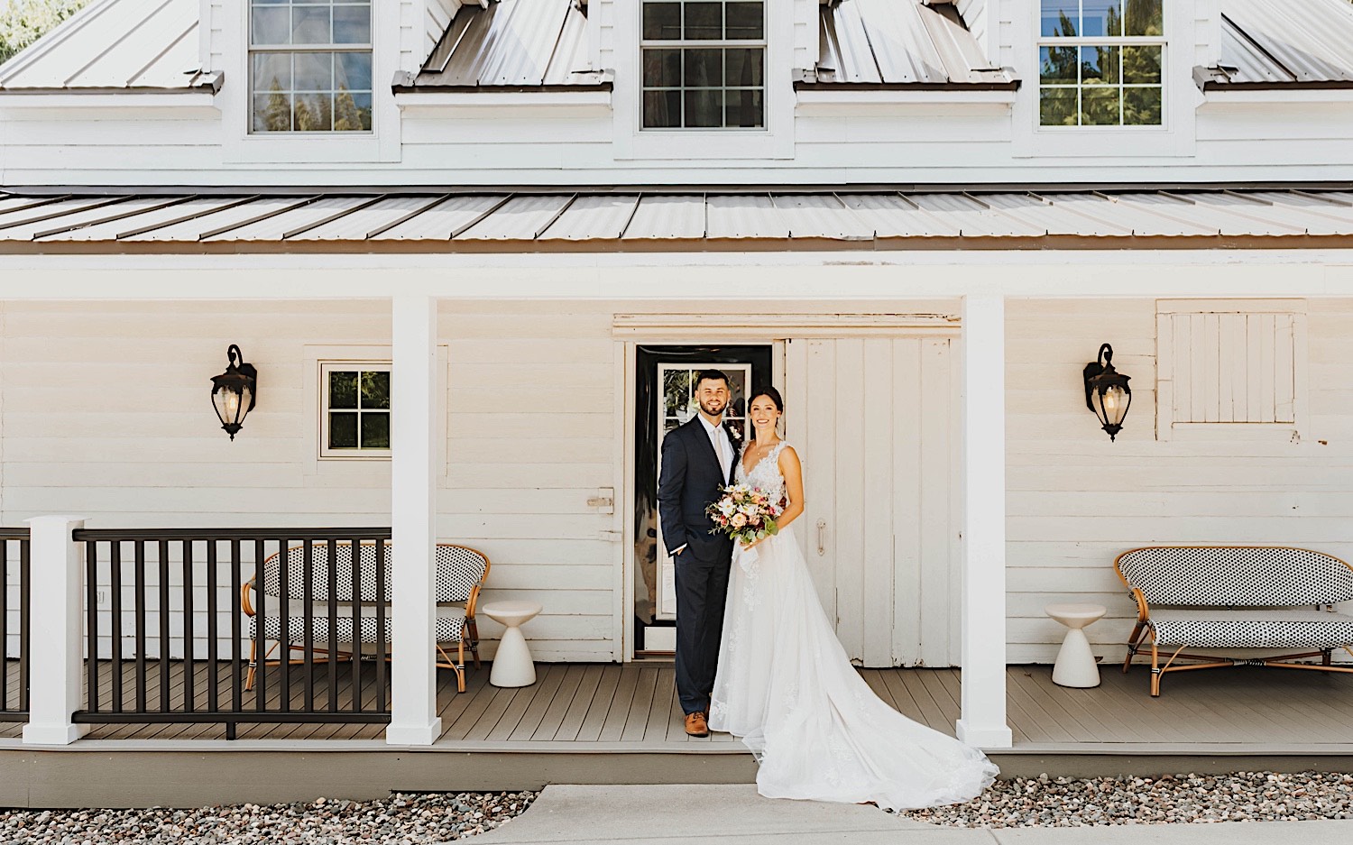 A bride and groom stand outside of a building of their wedding venue Legacy Hill Farms in Minnesota, they smile at the camera while holding one another A bride and groom stand outside of a building of their wedding venue Legacy Hill Farms in Minnesota, they smile at the camera while holding one another