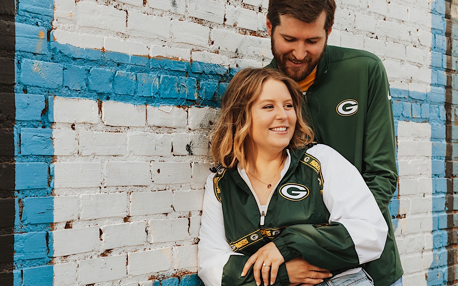 A woman smiles as a man hugs her from behind as the two lean against a brick wall while taking engagement photos in downtown Minneapolis