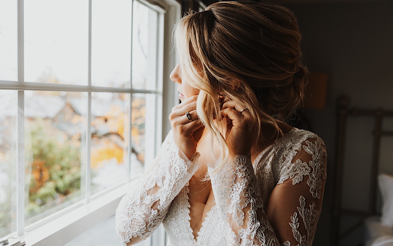 A bride in her wedding dress looks out of a window while putting on her earrings