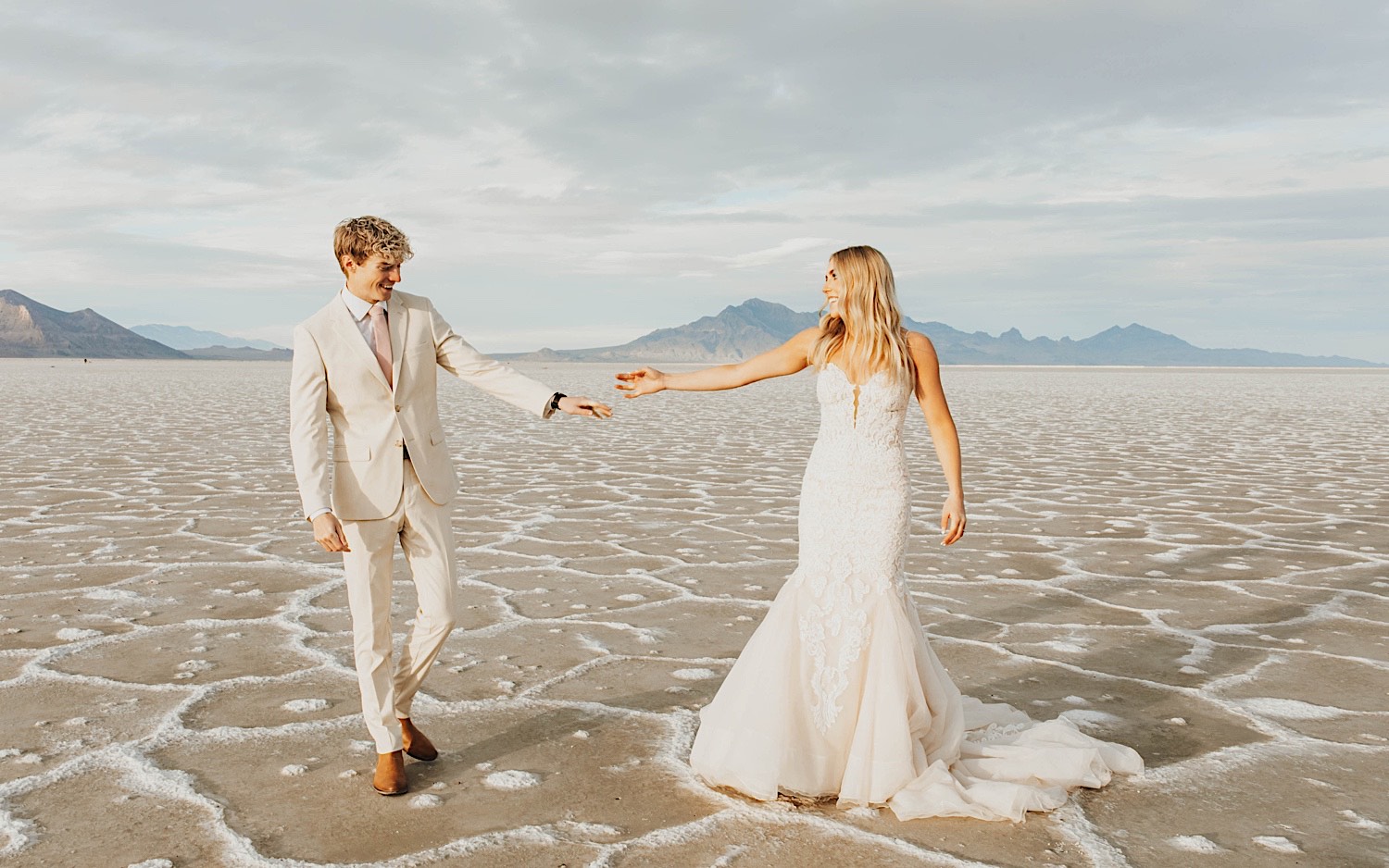 At their elopement in the Utah Salt Flats, a bride and groom smile at one another and reach their hands towards one another while walking towards the camera