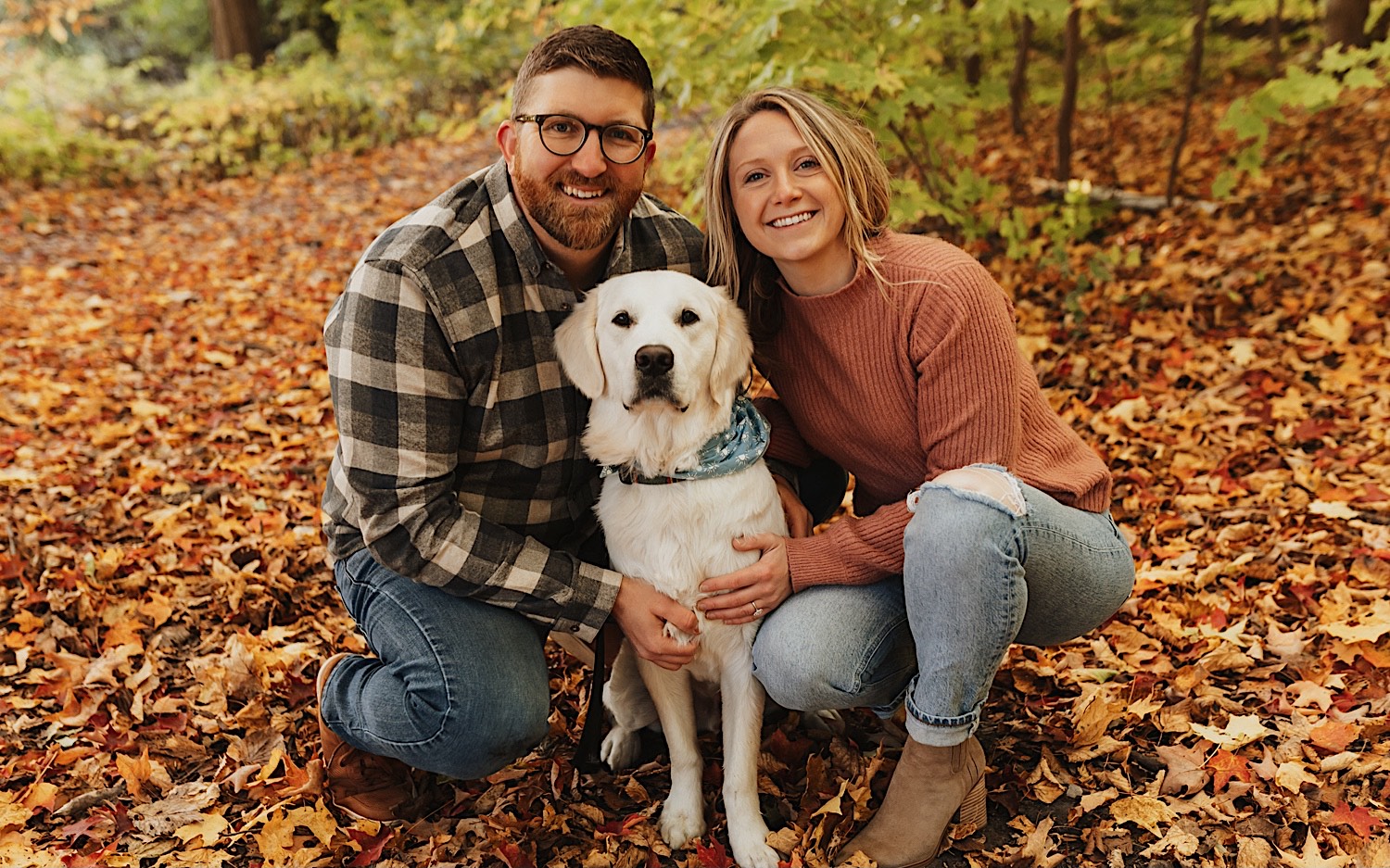 During their fall engagement session in Winona a couple squat down next to their dog who is sitting in between them, the three of them look at the camera and the man and woman smile, the ground around them is covered in leaves