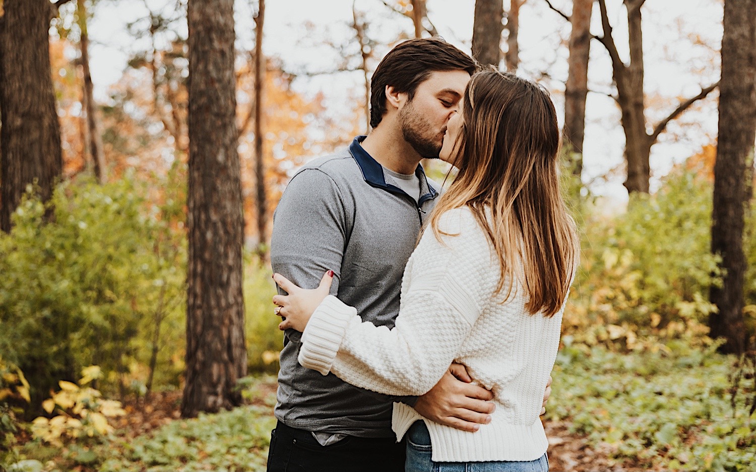 A couple kiss one another while in a forest of Theodore Wirth Park in Minneapolis during their engagement session
