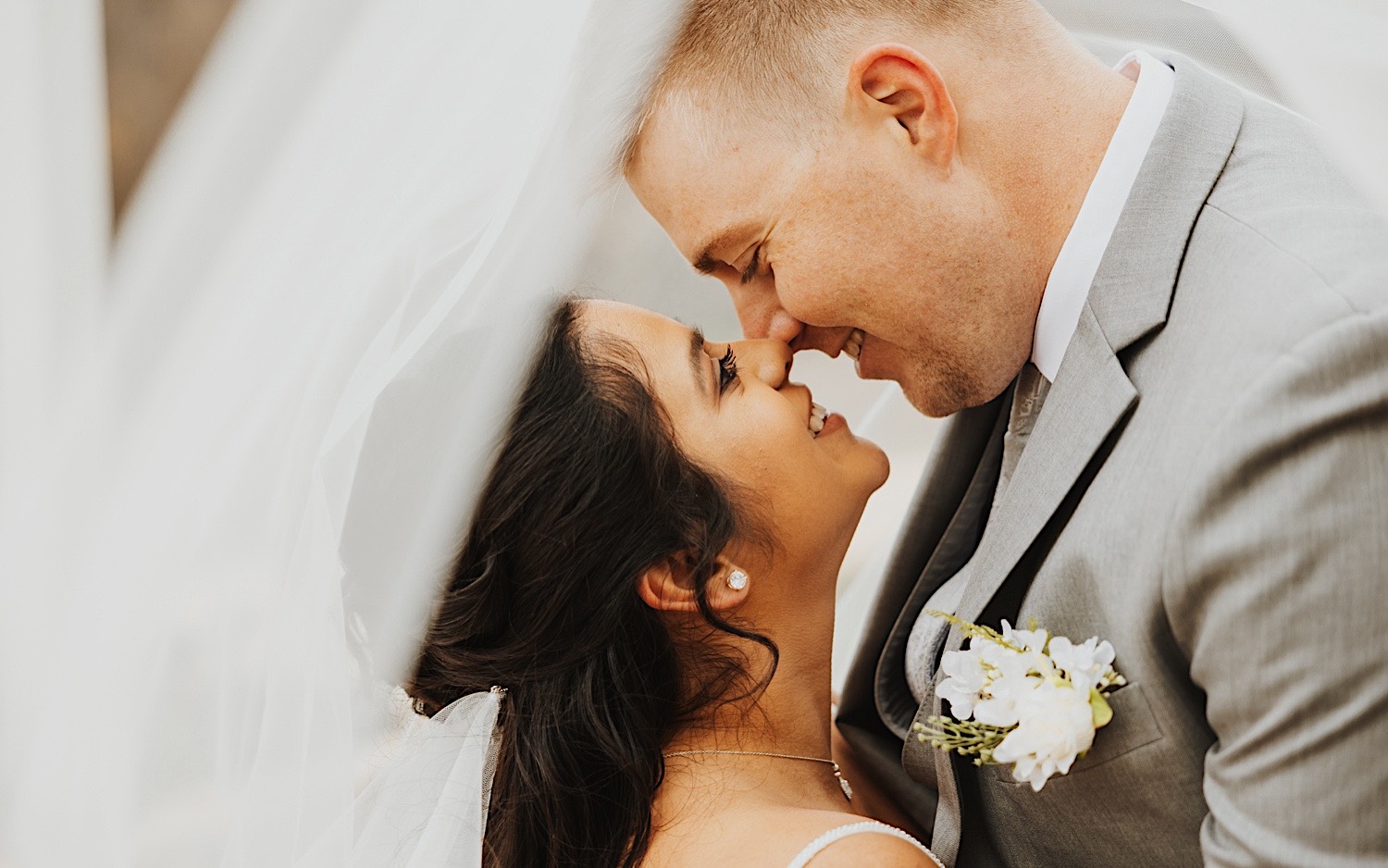 A bride and groom smile at each other before kissing underneath the bride's veil