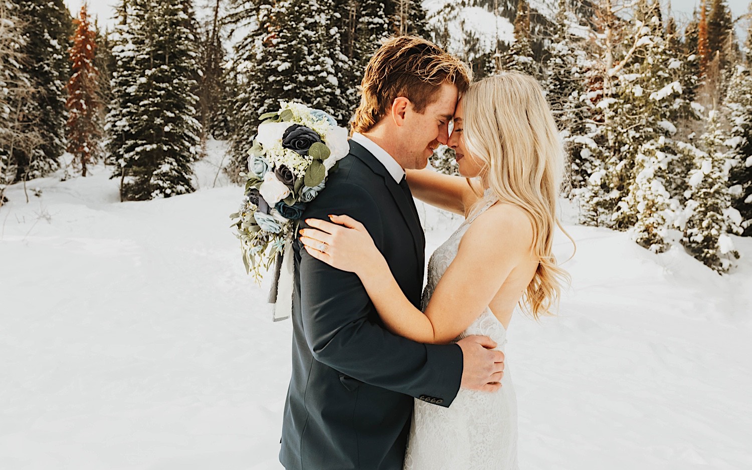 In a snow covered forest near Salt Lake City, a bride and groom smile at one another while touching their foreheads together during their elopement