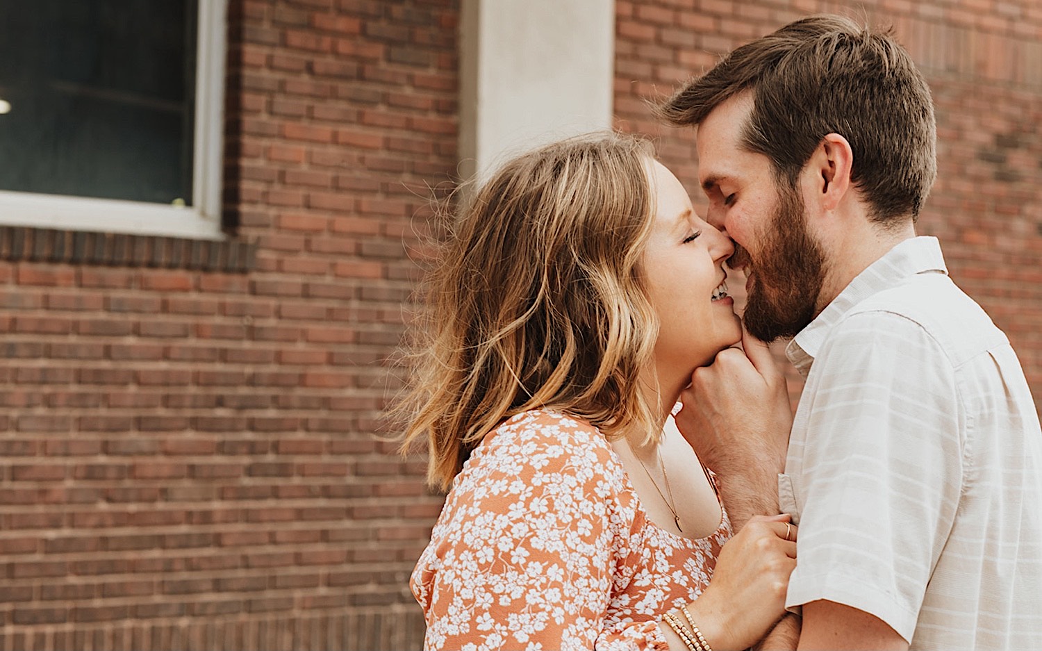 While taking engagement photos in downtown Minneapolis a couple smile as they lean in to kiss one another while in front of a brick wall