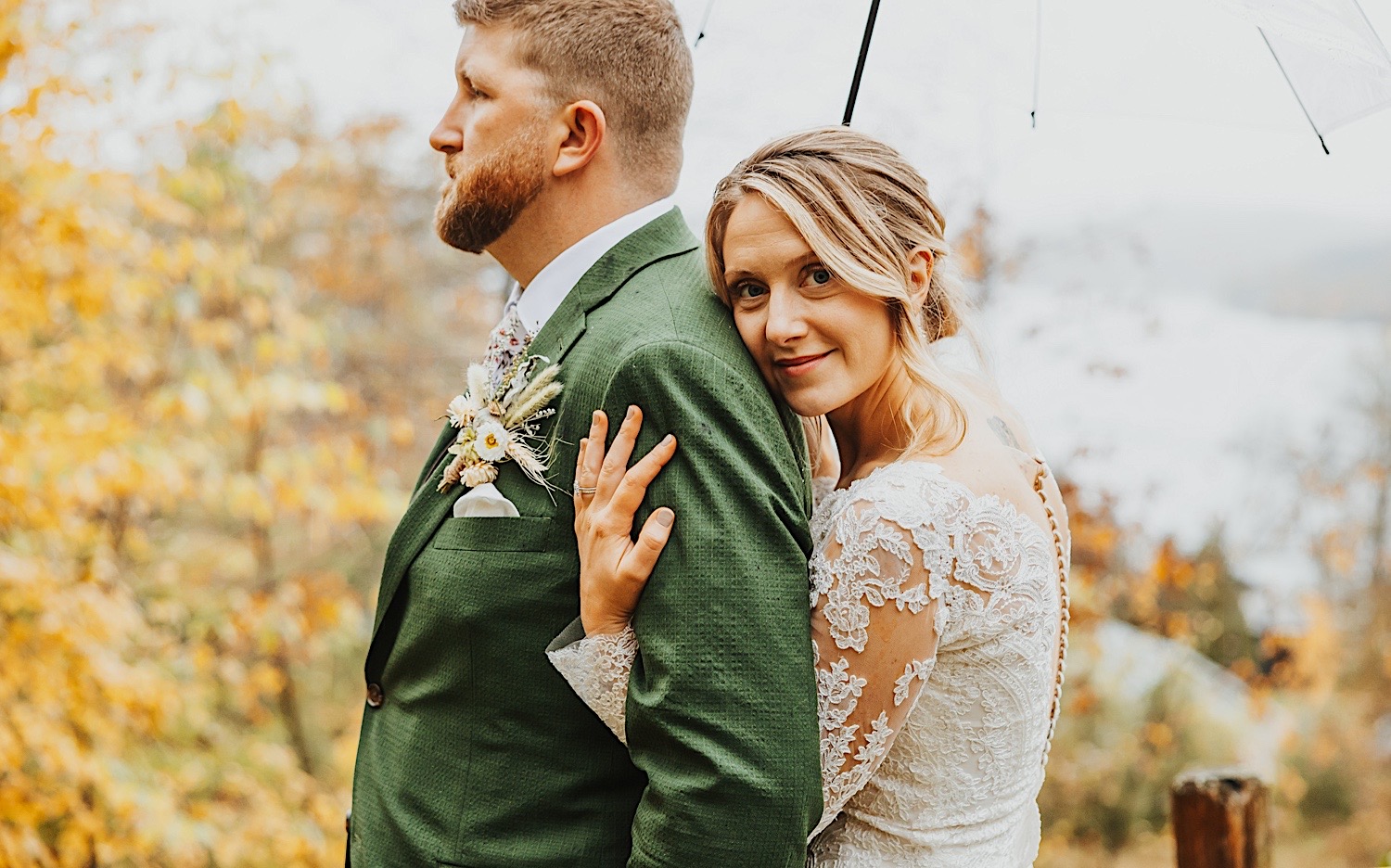 A bride looks at the camera while hugging the groom from behind while underneath an umbrella, behind them are trees with yellow leaves