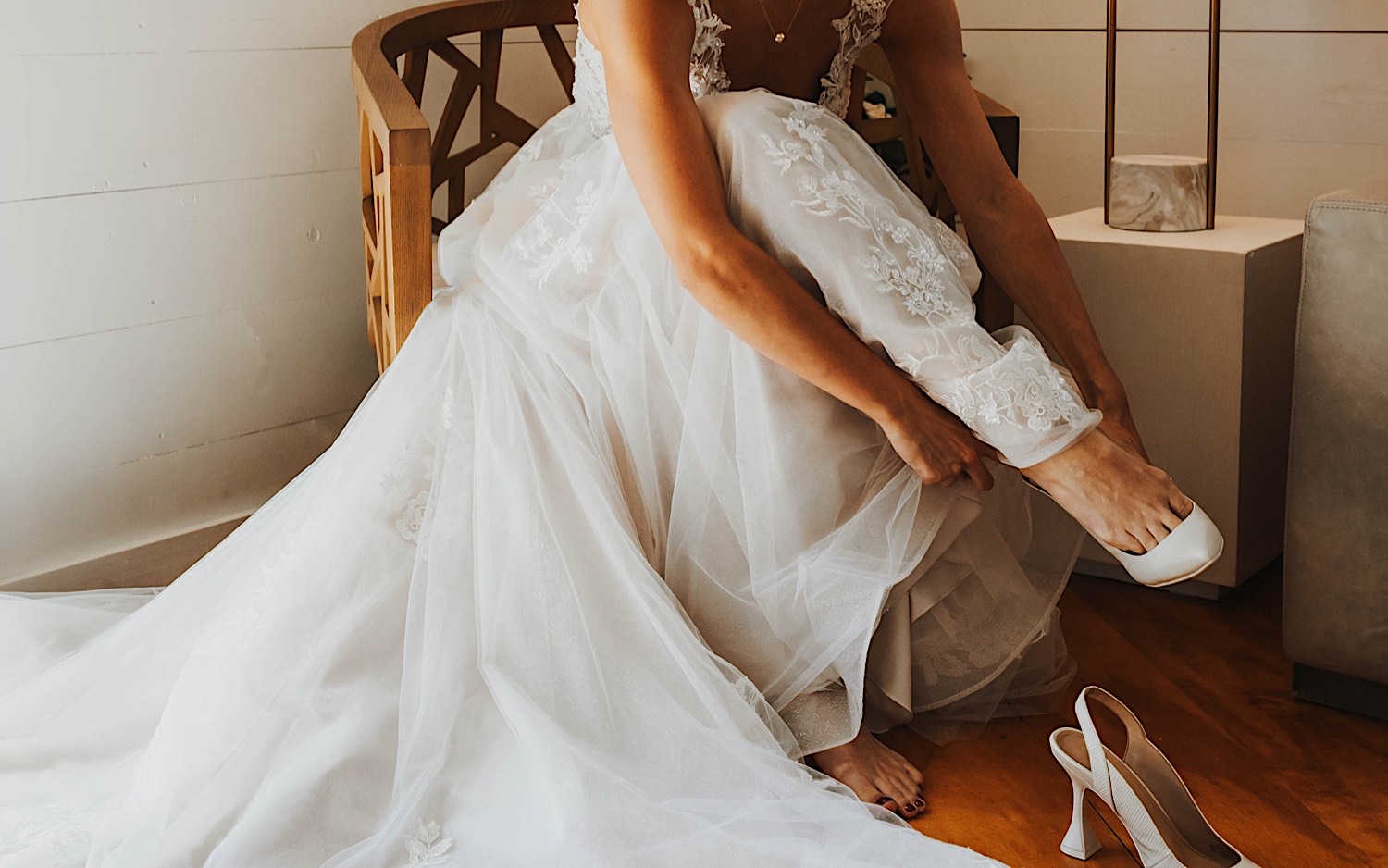 A bride sits in a chair in her wedding dress and puts on her shoes while in the getting ready space at Legacy Hill Farm