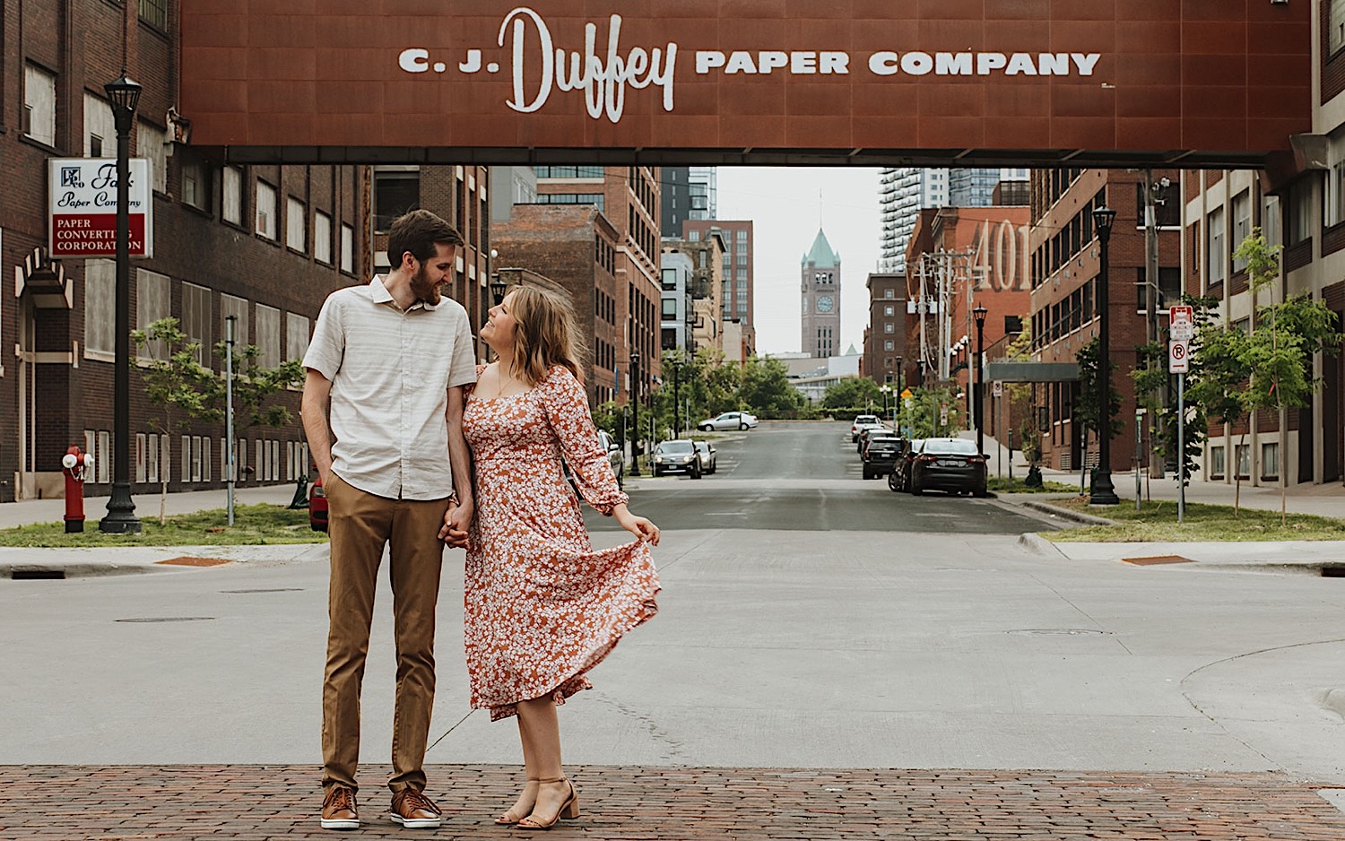 A couple stand side by side and hold hands while in front of a skyway with the Duffey Paper Company logo on it, the two are taking engagement photos in downtown Minneapolis