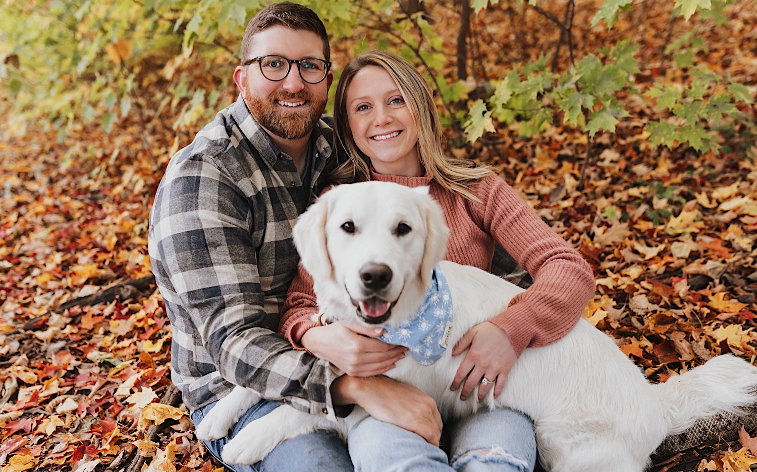 A couple sit on the ground next to one another with leaves all around them and smile at the camera during their fall engagement session in Winona, their dog is laying across their laps and is also looking at the camera