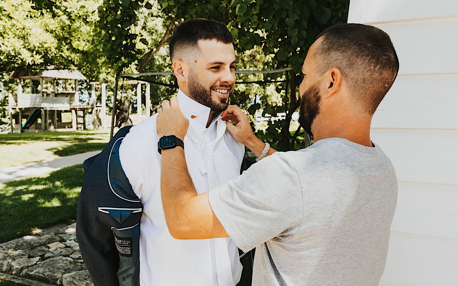 A groom smiles while standing outside as a friend helps adjust his shirt collar before his wedding at Legacy Hill Farm