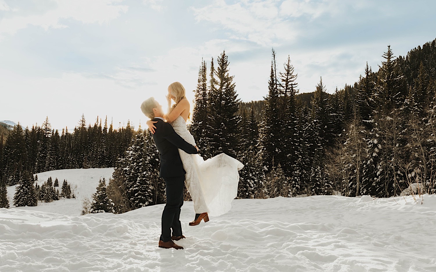 During their elopement in a snow covered park near Salt Lake City a groom lifts a bride into the air as they smile at one another while the sun sets behind them