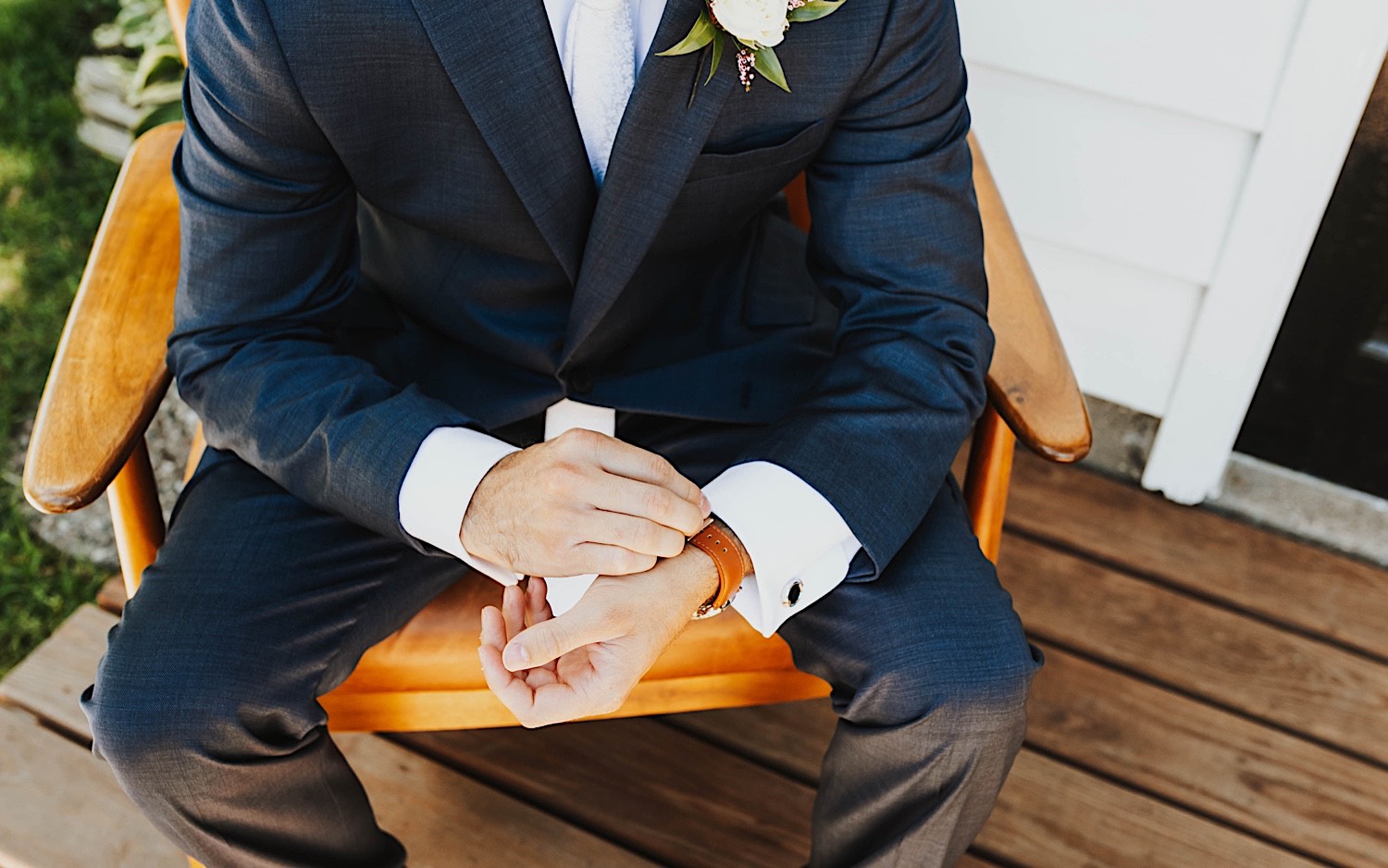 A groom sits in a chair on a front patio and adjusts his suit cuffs