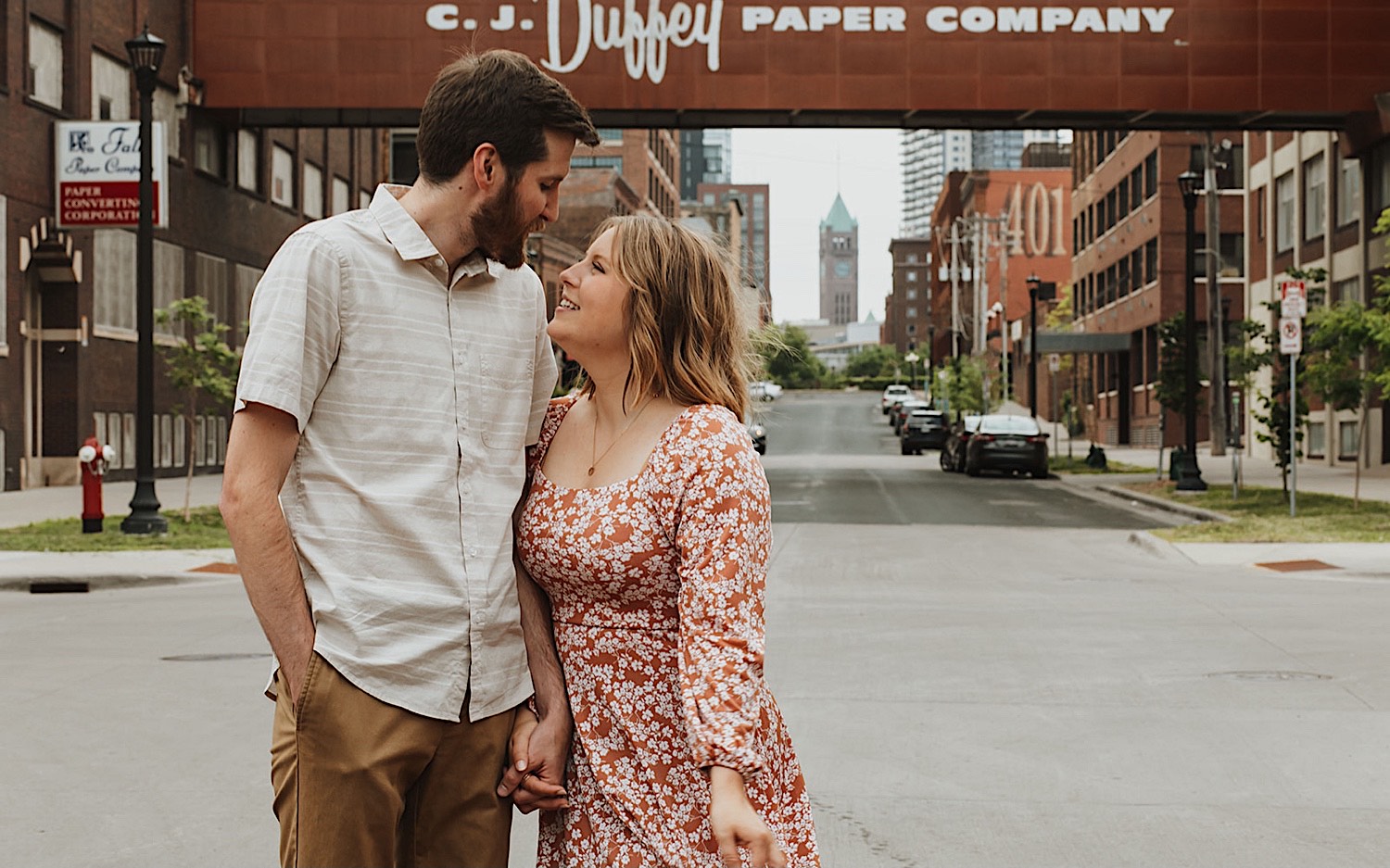 While taking engagement photos in downtown Minneapolis a couple hold hands and smile at one another while in front of the skyway of the Duffey Paper Company