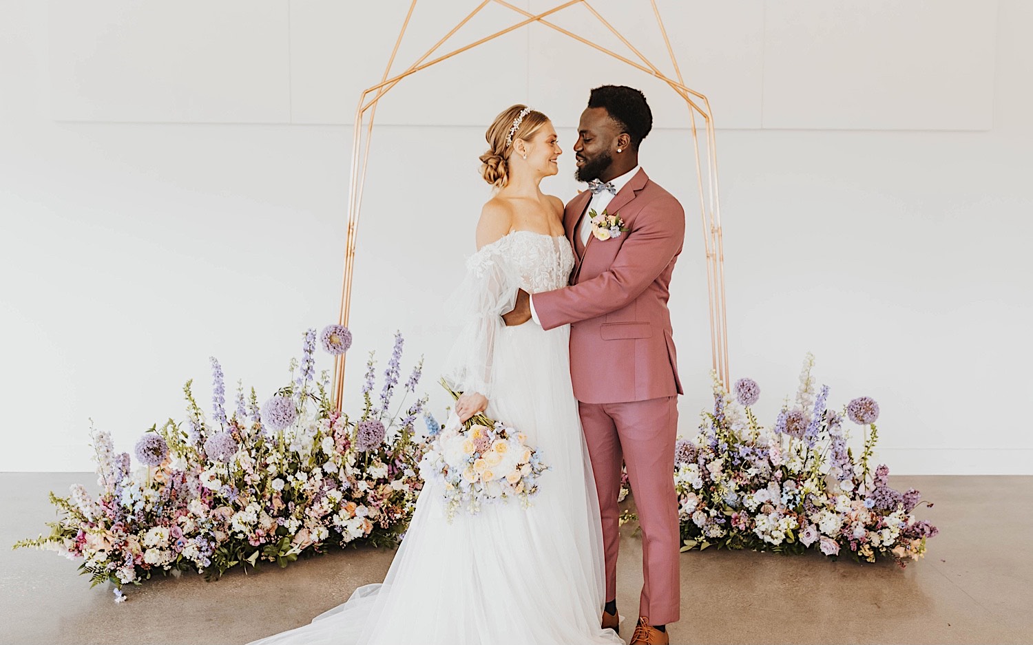 A bride and groom embrace in front of flowers and a wedding archway during their wedding ceremony at The Aisling