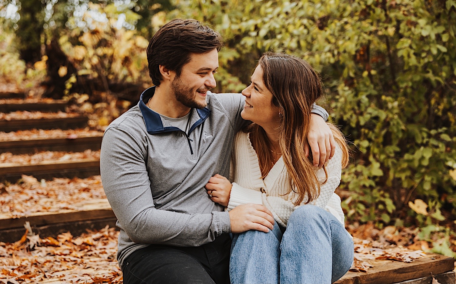 A couple smile at one another as they sit side by side on a wooden staircase inside of Theodore Wirth Park in Minneapolis during their engagement session