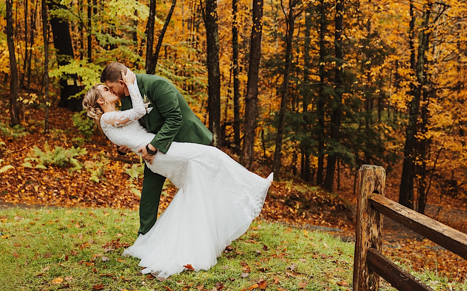 A bride and groom kiss one another while in the grass next to a forest of yellow trees in the fall