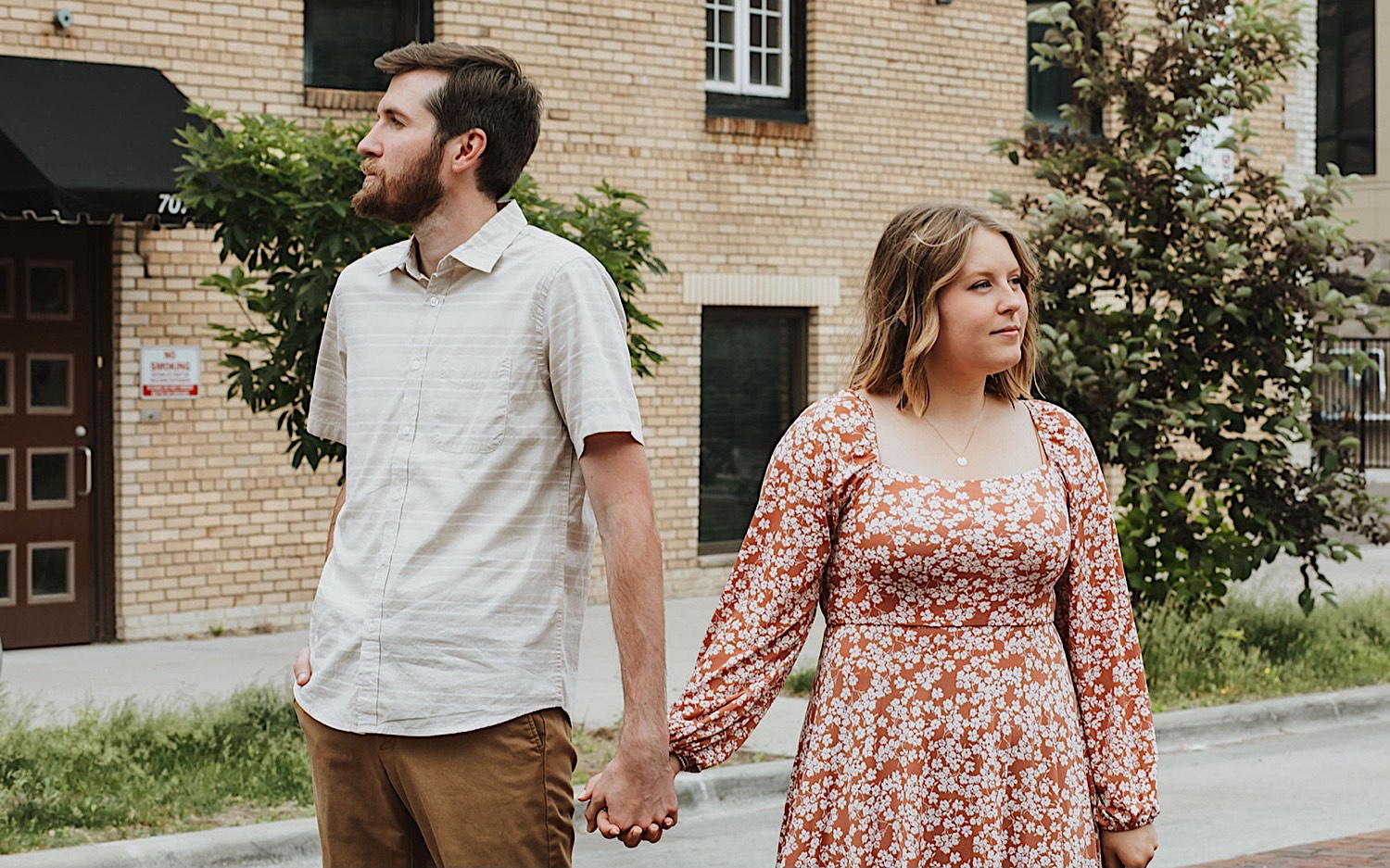 While taking engagement photos in downtown Minneapolis, a couple hold hands while facing and looking away from one another in front of a brick building