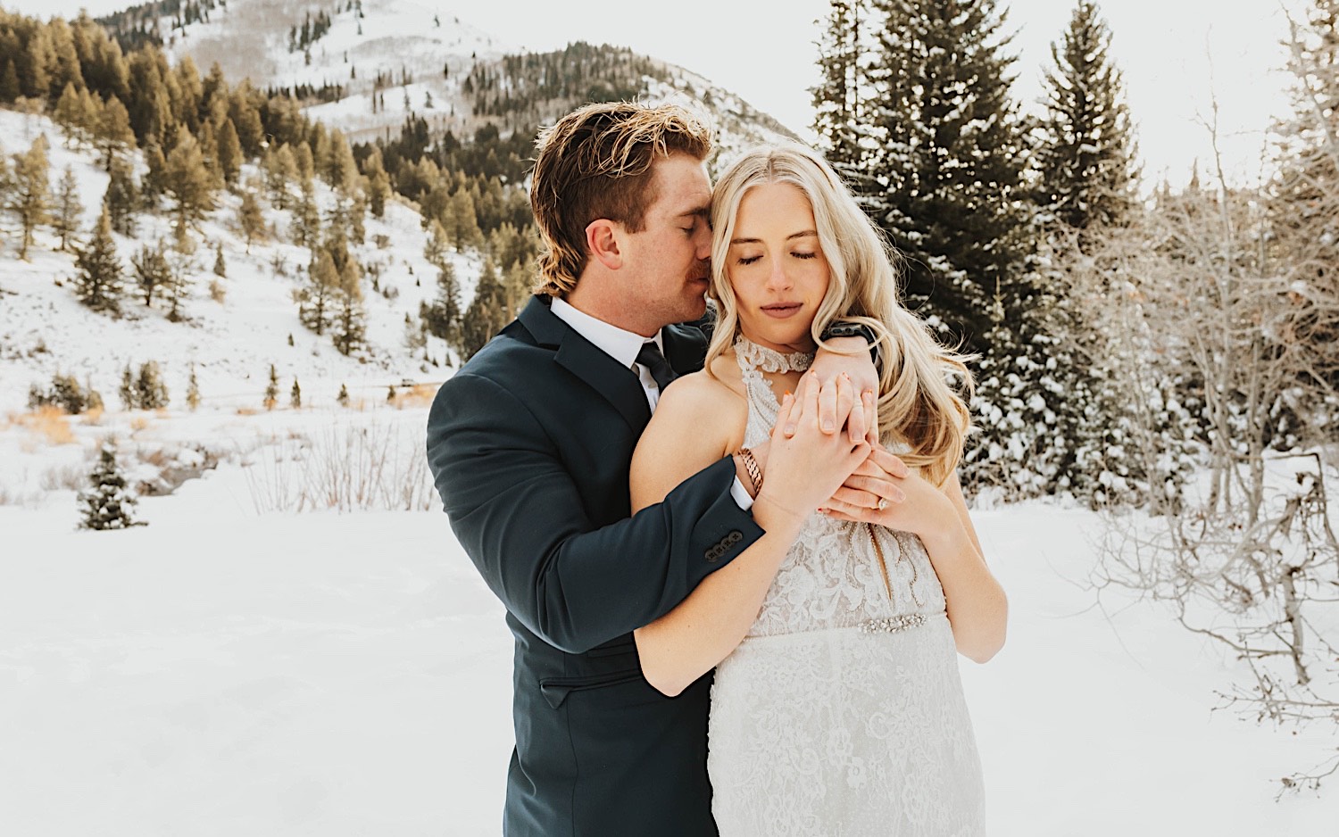 In a snow covered forest in a mountain near Salt Lake City a bride closes her eyes while a groom stands behind her and is about to kiss her cheek