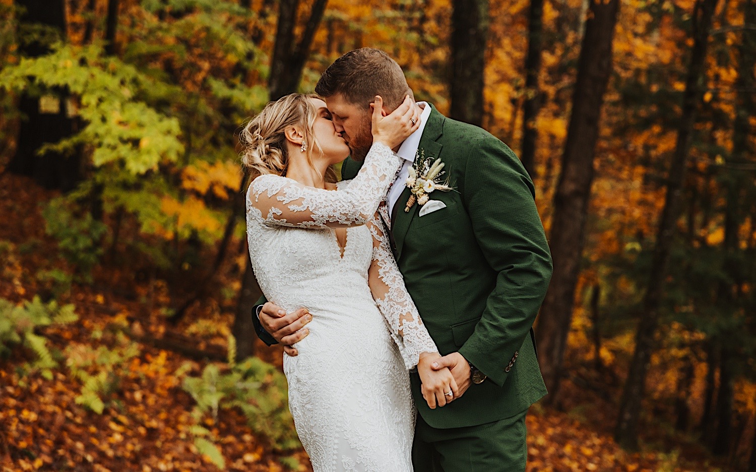 A bride and groom kiss one another while in front of a forest, it is the fall so the leaves are different shades of yellow
