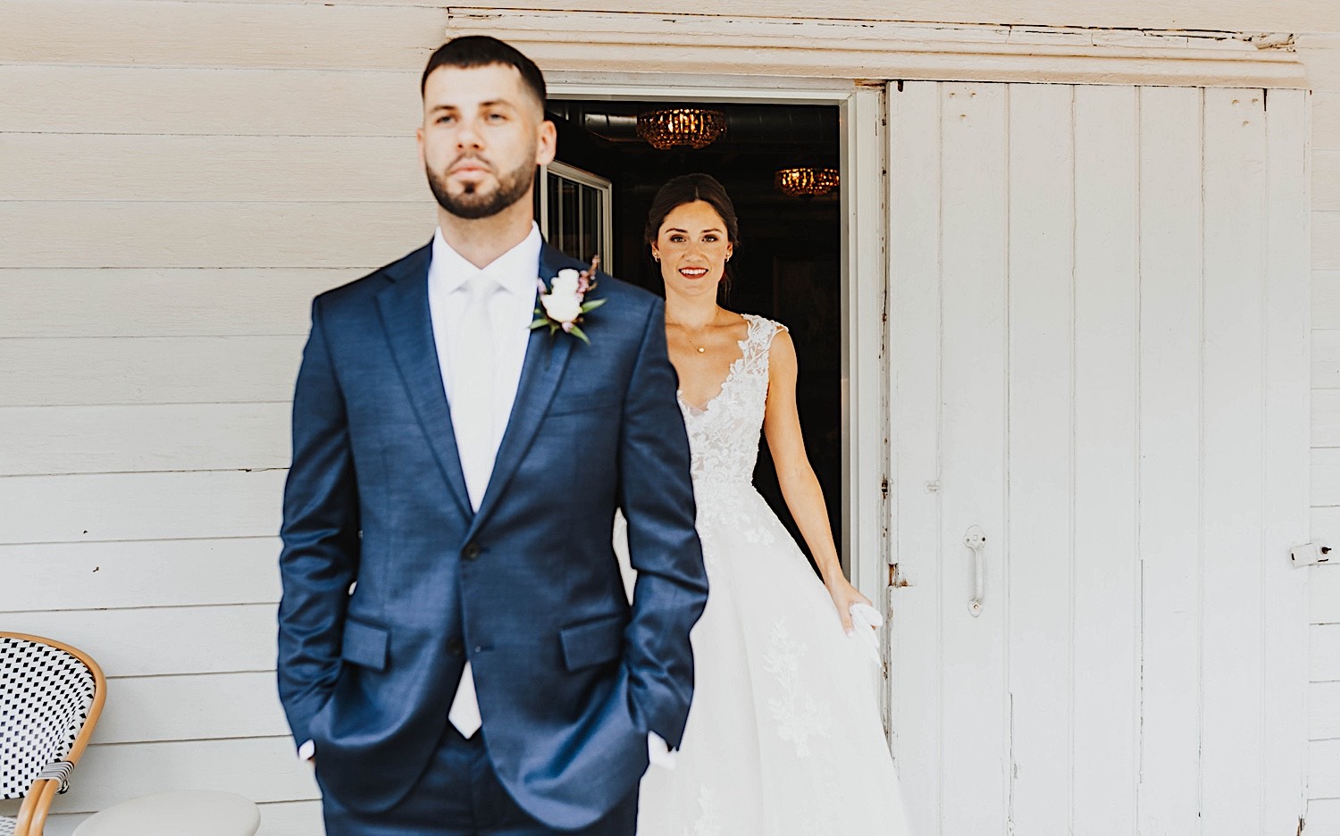 A groom faces the camera while standing outside of a white building, behind him in the doorway is the bride about to tap him on the shoulder for a first look on their wedding day at Legacy Hill Farm