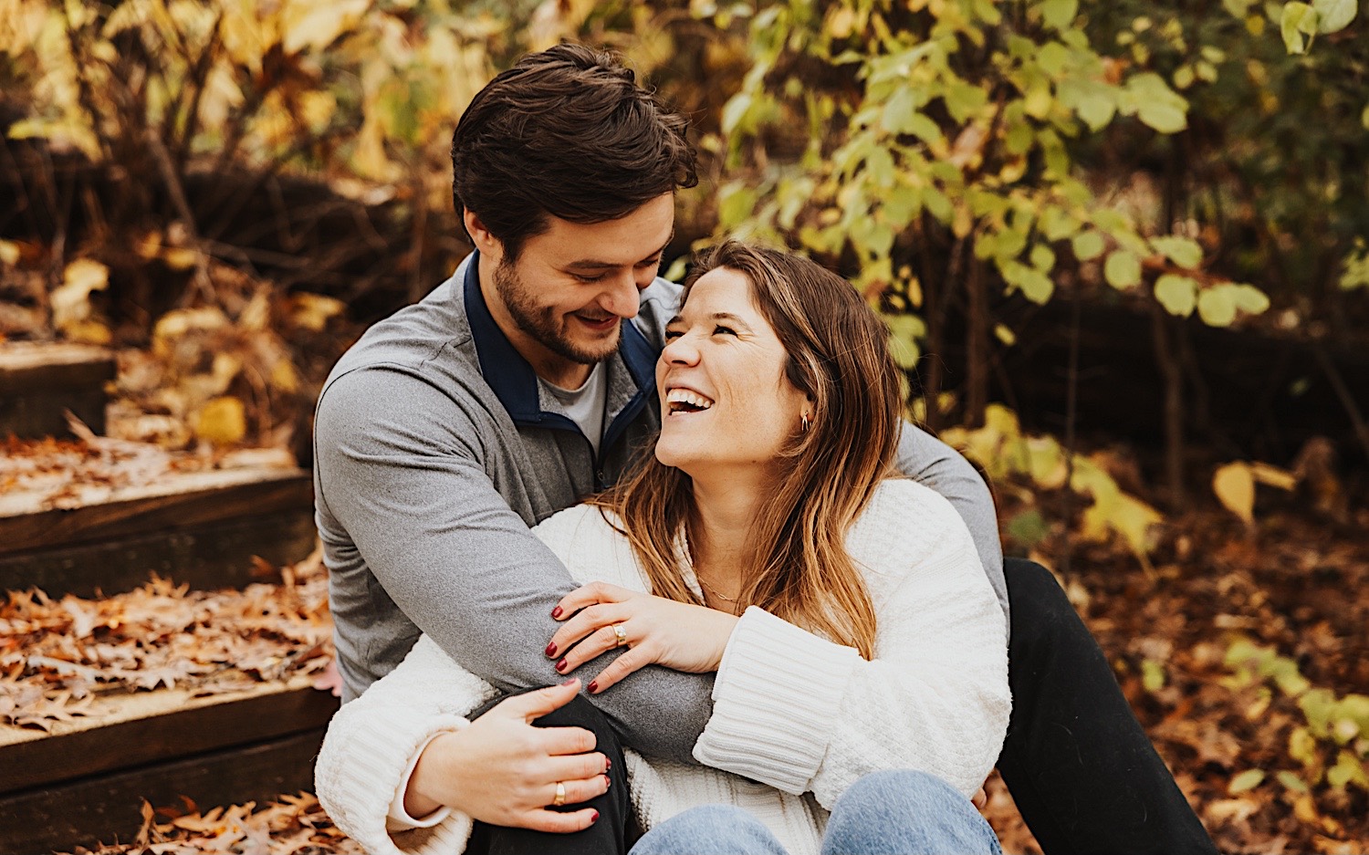 A couple laugh as they sit together on a wooden staircase inside of Theodore Wirth Park of Minneapolis during their engagement session