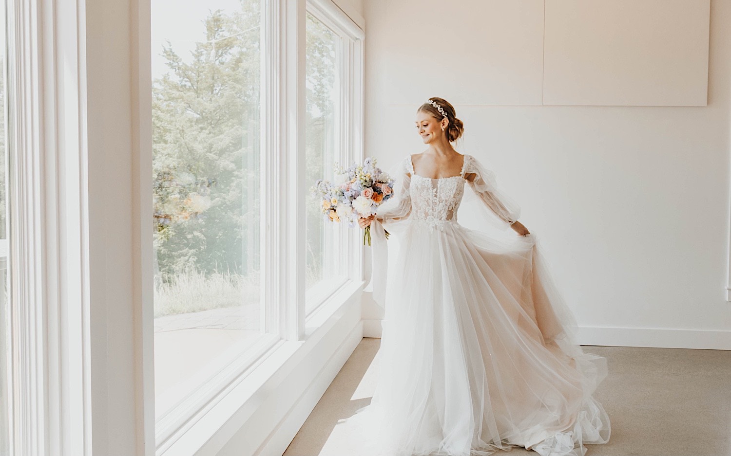A bride stands in front of a window holding her bouquet and playing with her wedding dress during her wedding day at The Aisling