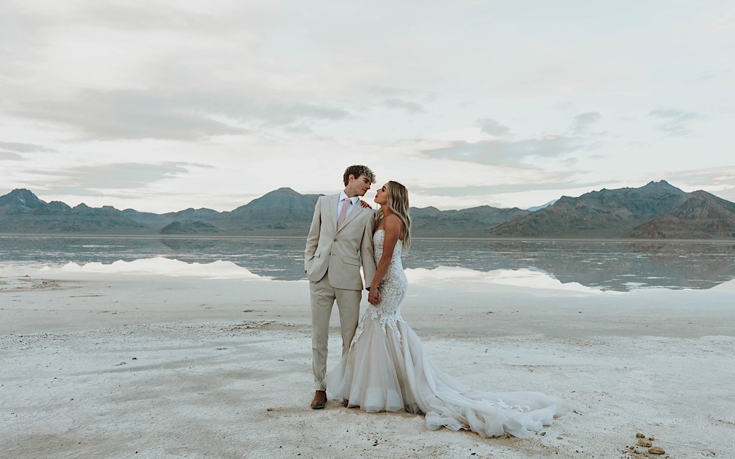During an elopement at the Utah Salt Flats near Salt Lake City, a bride and groom stand next to one another and are about to kiss with the mountains and salt flats behind them