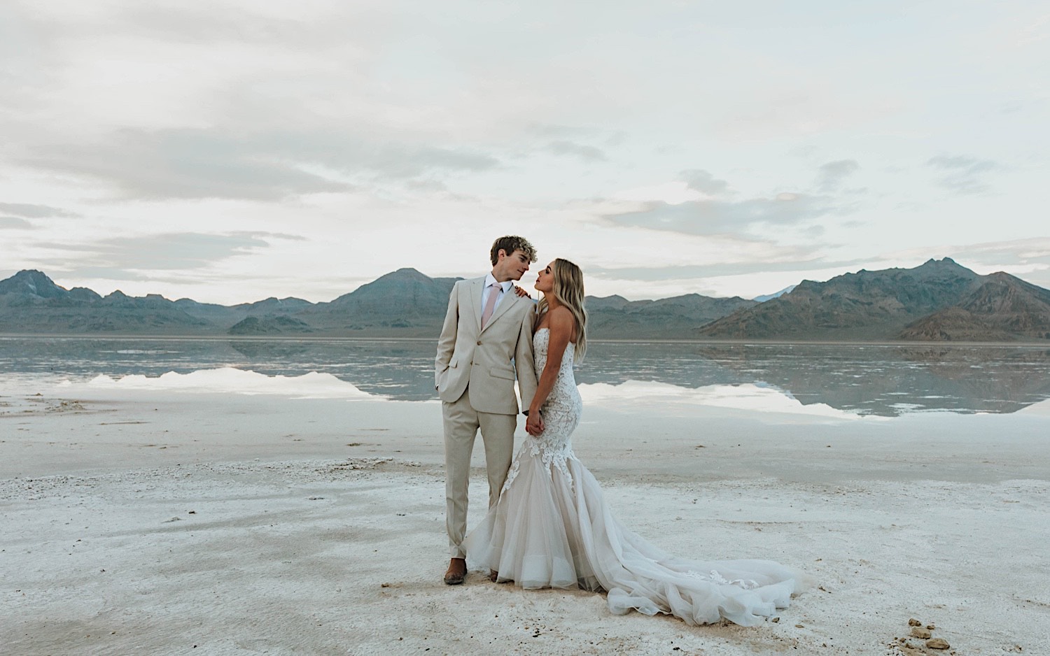During an elopement at the Utah Salt Flats near Salt Lake City, a bride and groom stand next to one another and are about to kiss with the mountains and salt flats behind them During an elopement at the Utah Salt Flats near Salt Lake City, a bride and groom stand next to one another and are about to kiss with the mountains and salt flats behind them