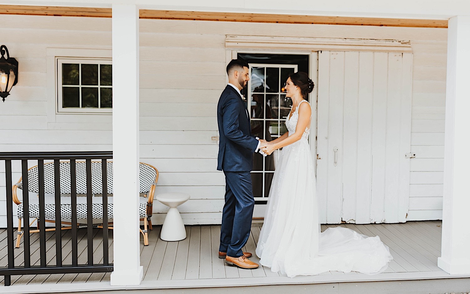 A bride and groom hold hands while facing one another on a front patio of a white building of the wedding venue Legacy Hill Farm