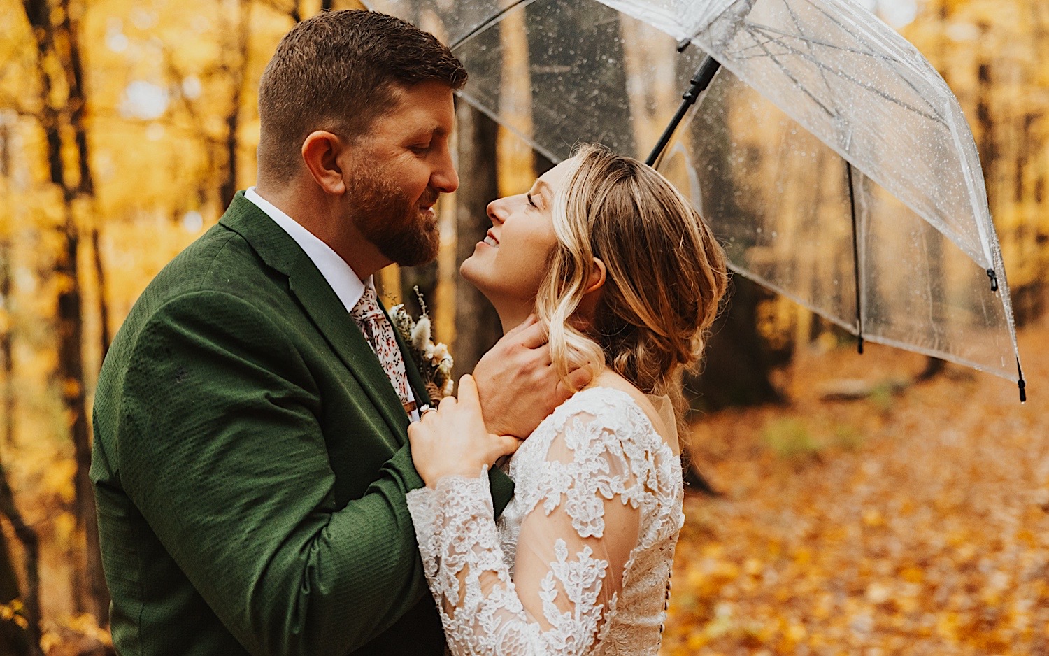 A bride and groom smile at one another while under an umbrella in a leaf covered forest during their wedding day at Lake Bomoseen Lodge in Vermont
