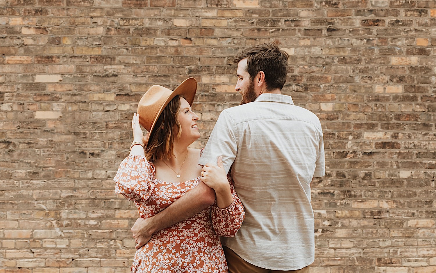 While in front of a brick wall a couple stand side by side and look at one another while facing opposite directions 