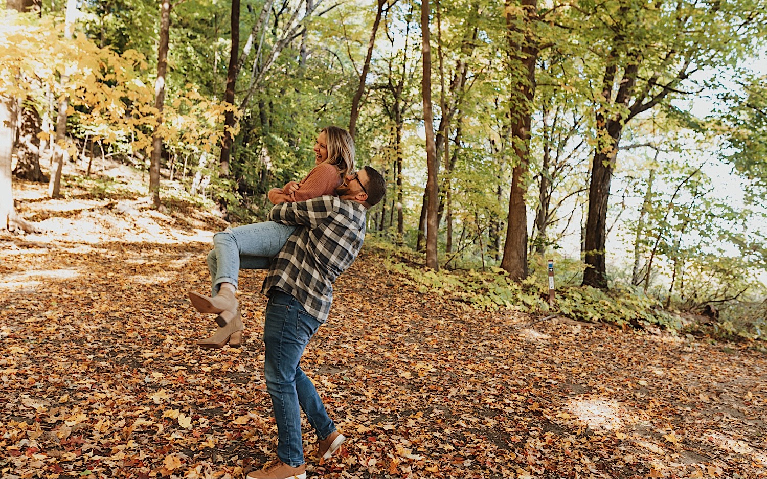 During their fall engagement session in Winona a woman laughs as a man lifts her in the air from behind and spins her around, they're on a trail in the forest covered in leaves